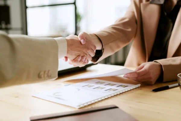 Close-up of two business professionals shaking hands over a wooden desk with financial documents.