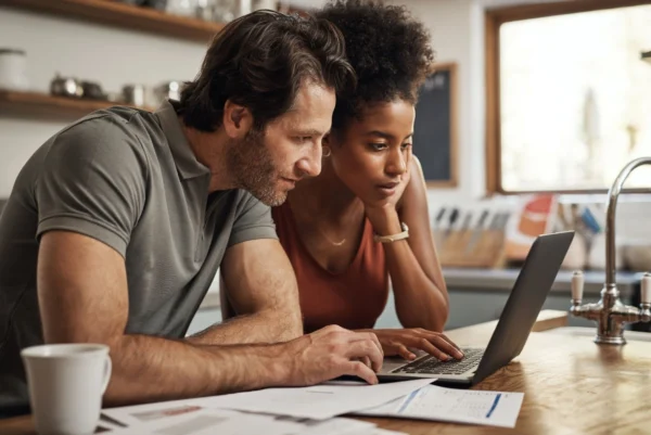 A young couple sitting in front of a laptop on the kitchen bench along with various pieces of paperwork and a cup of coffee.